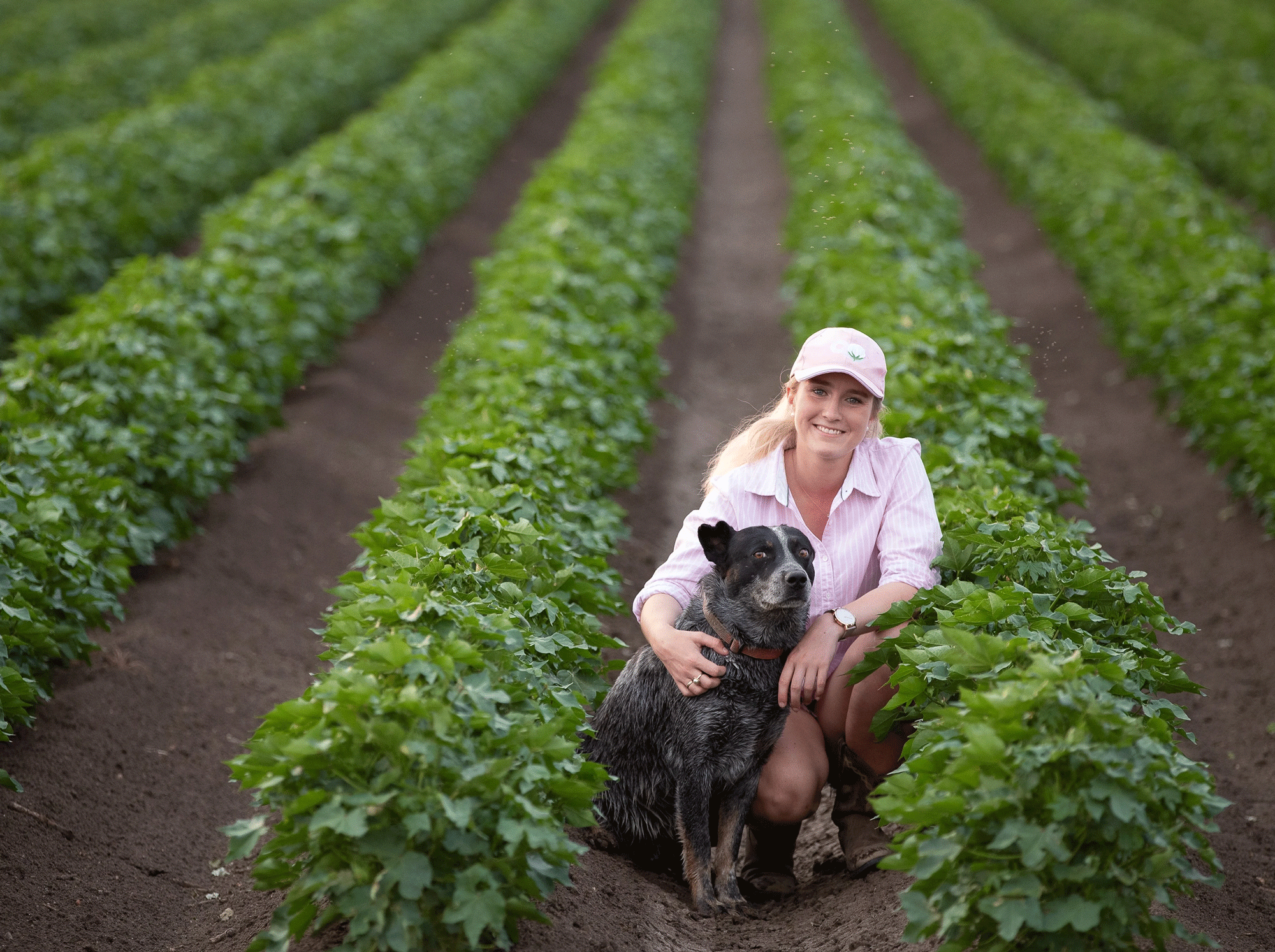 A female agricultural worker kneeling in a green crop field with a cattle dog, smiling at the camera.