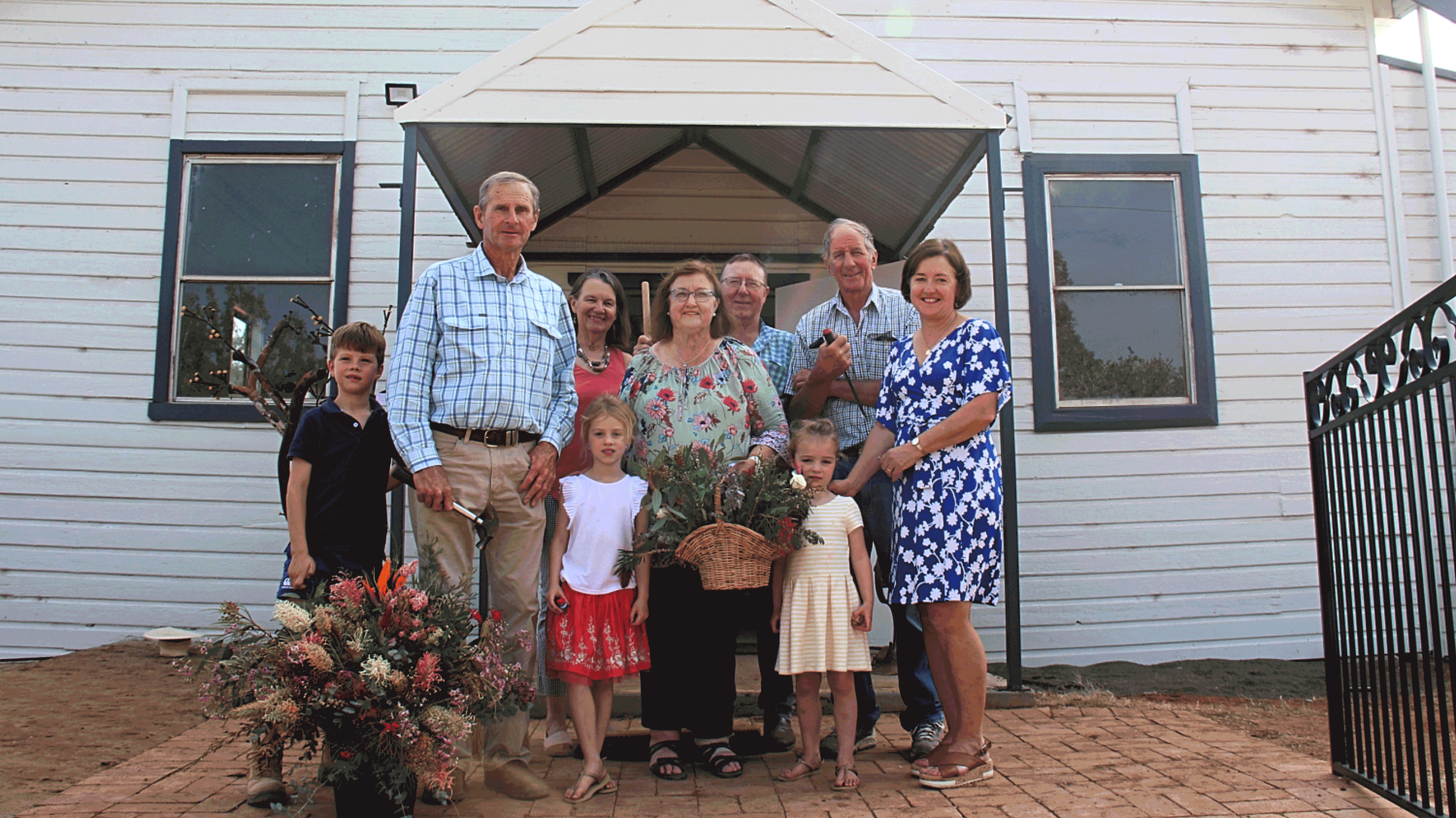 A group of adults and children standing in front of a white building, smiling and holding flowers and baskets.
