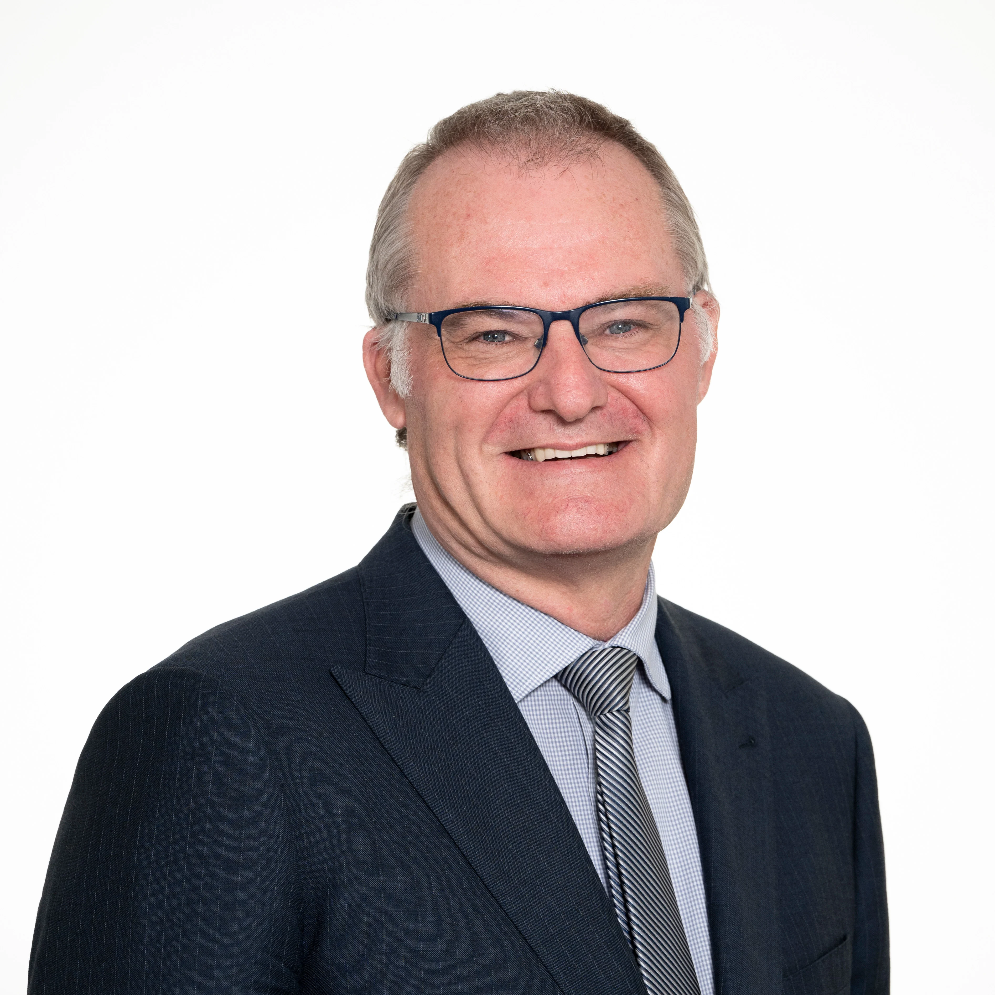 Headshot of Chief Executive Brock Gilmour, smiling and wearing glasses, a dark suit, and a grey patterned shirt with a tie.