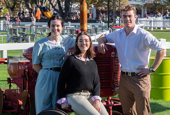 Three young adults posing beside a vintage red tractor at an outdoor event.