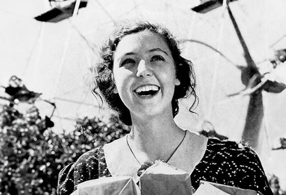 Black and white photo of a smiling woman standing in front of a ferris wheel. 