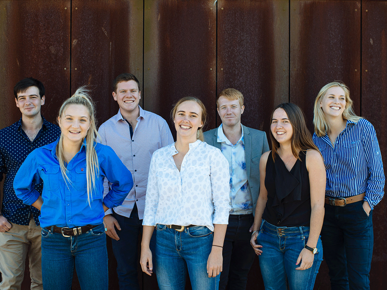 Seven smiling agricultural scholars in casual smart clothing stand outdoors against a rust-coloured wall.