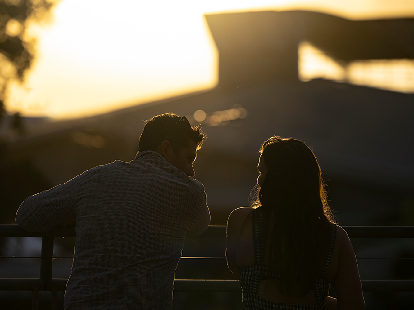 A man and woman facing back to the camera, leaning on a railing at sunset, silhouetted against a golden sky.