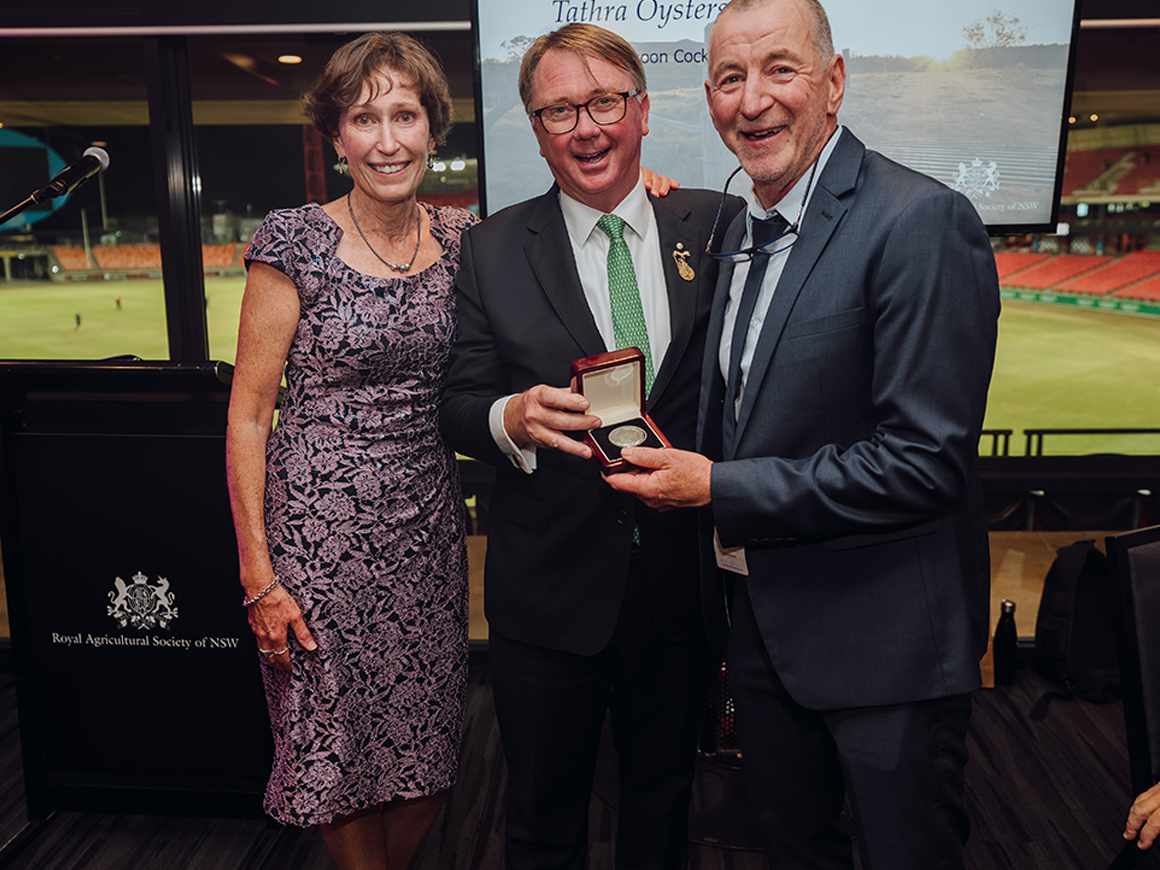 Two men and a woman smiling as Tathra Oysters receive the 18th Annual President’s Medal on stage.