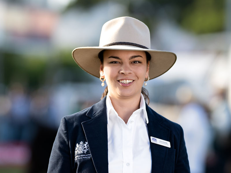 Headshot of 2026 RAS Rural Achiever Anna Sutcliffe, a young woman with dark brown hair with hair tied back and earrings, wearing a wide-brim hat in a navy blazer.