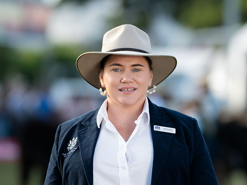 Headshot of 2026 RAS Rural Achiever Tara Kennedy, a young woman with hair pulled back with hoop earings wearing wide-brim hat in a navy blazer.
