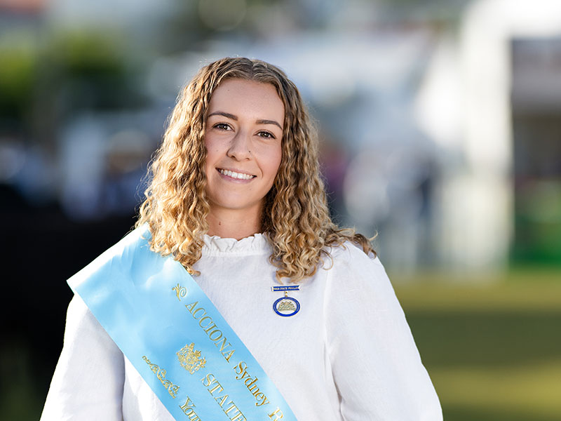 Headshot of 2026 Young Woman finalist Bianca Evans, a blonde woman with curly hair, wearing a white shirt and a light blue sash.