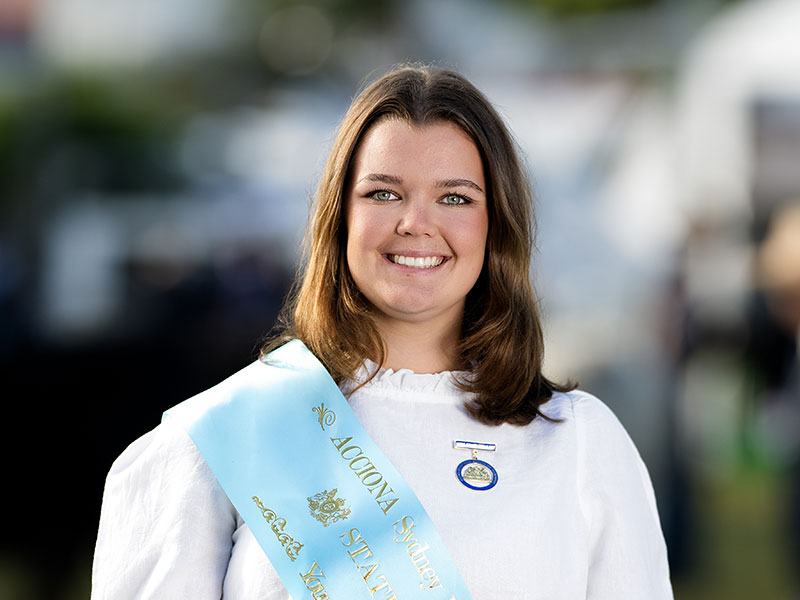 Headshot of 2026 Young Woman finalist Eva Price, a light brunette woman with shoulder-length hair, wearing a white shirt and a light blue sash.