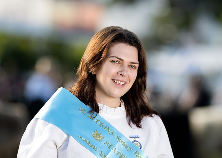 Headshot of 2026 Young Woman winner Louisa Gilmour, a woman with dark brown shoulder-length hair in a white shirt and light blue sash.