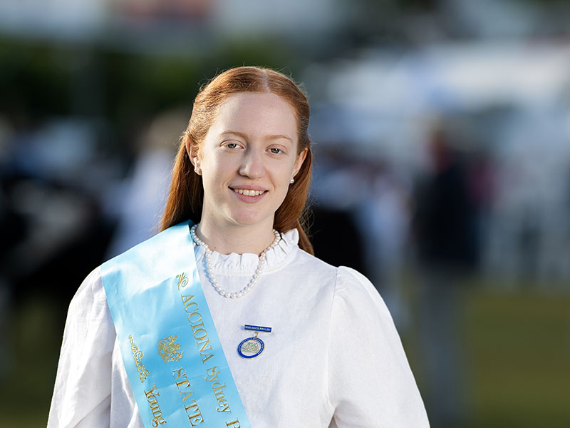 Headshot of 2026 Young Woman finalist Paige Sedgwick, a woman with ginger hair pulled back, wearing a white shirt and a light blue sash.