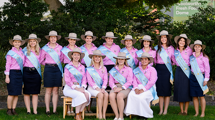 Group of young women in pink striped shirts, navy or white skirts, and hats, posing outdoors with light blue sashes for The Land Sydney Royal AgShows NSW Young Woman.
