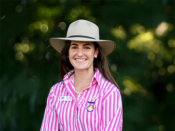 Headshot of 2025 finalist Brianna Pilon, a woman with long dark hair, wearing a wide-brimmed hat, a pearl necklace and earrings.