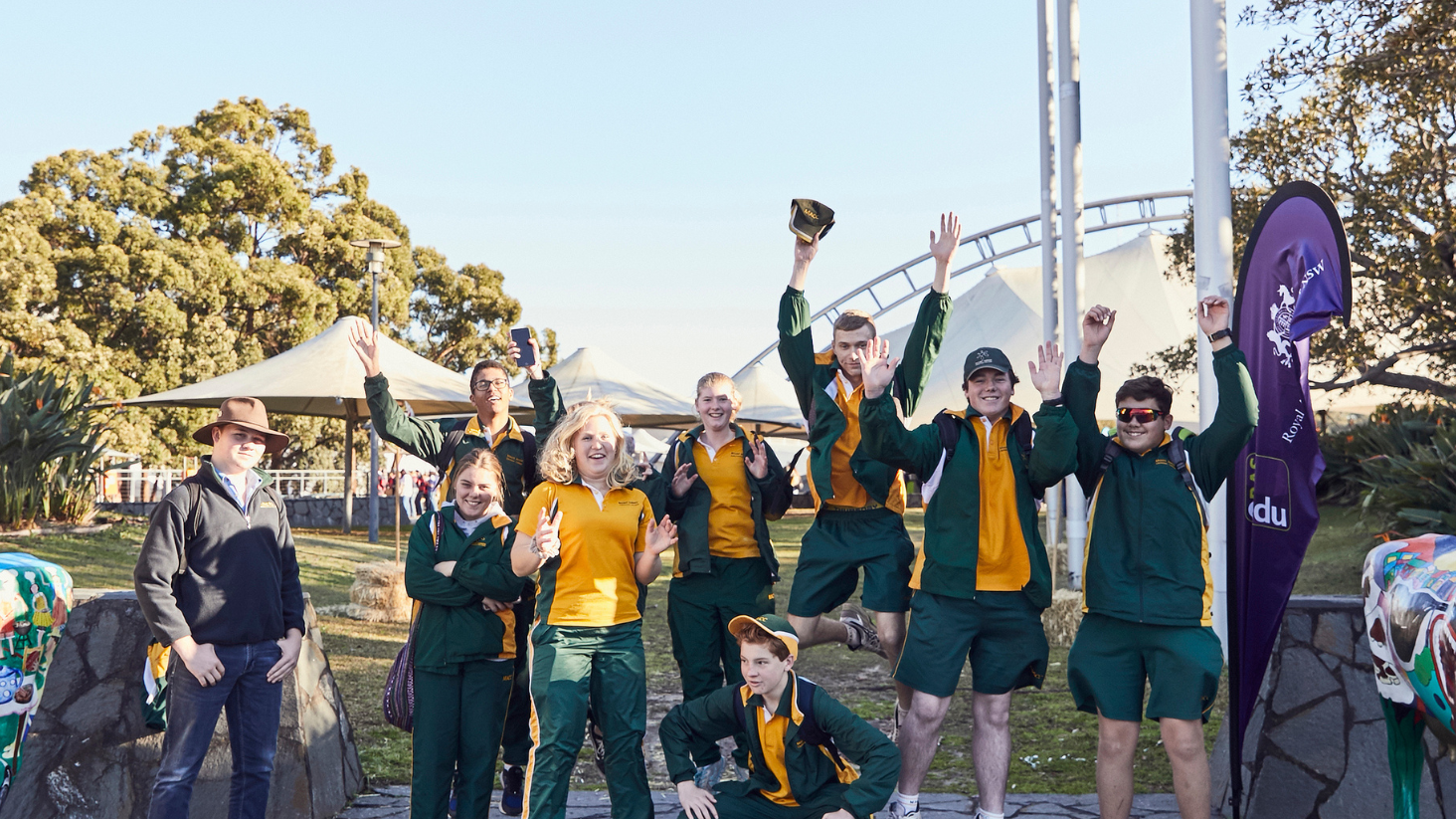 Group of students outdoors in school uniforms, smiling and jumping with excitement at an agricultural event.