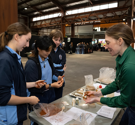 Three students in navy uniforms interact with a presenter demonstrating agricultural samples on a table.