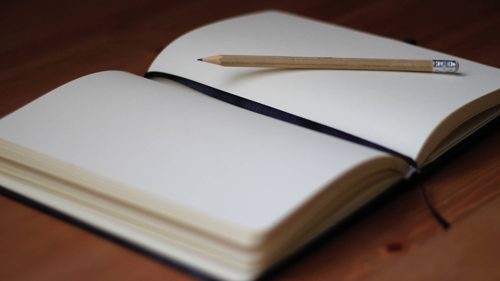 Open notebook with a pencil resting across blank pages on a wooden desk.