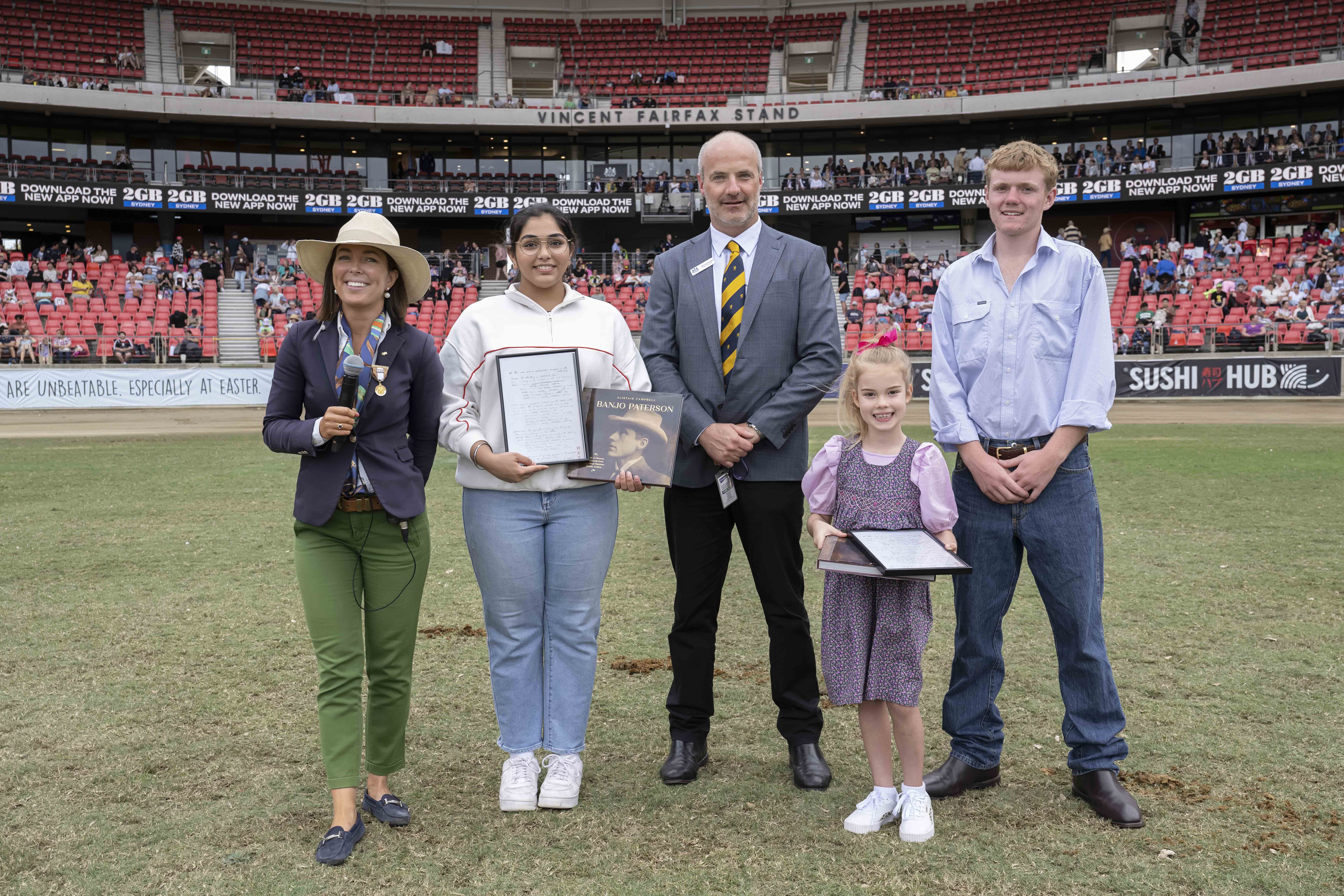 A group of five people standing in ENGIE Stadium, including the two 2024 RAS AB Paterson Bush Poetry Award Winners holding certificates.