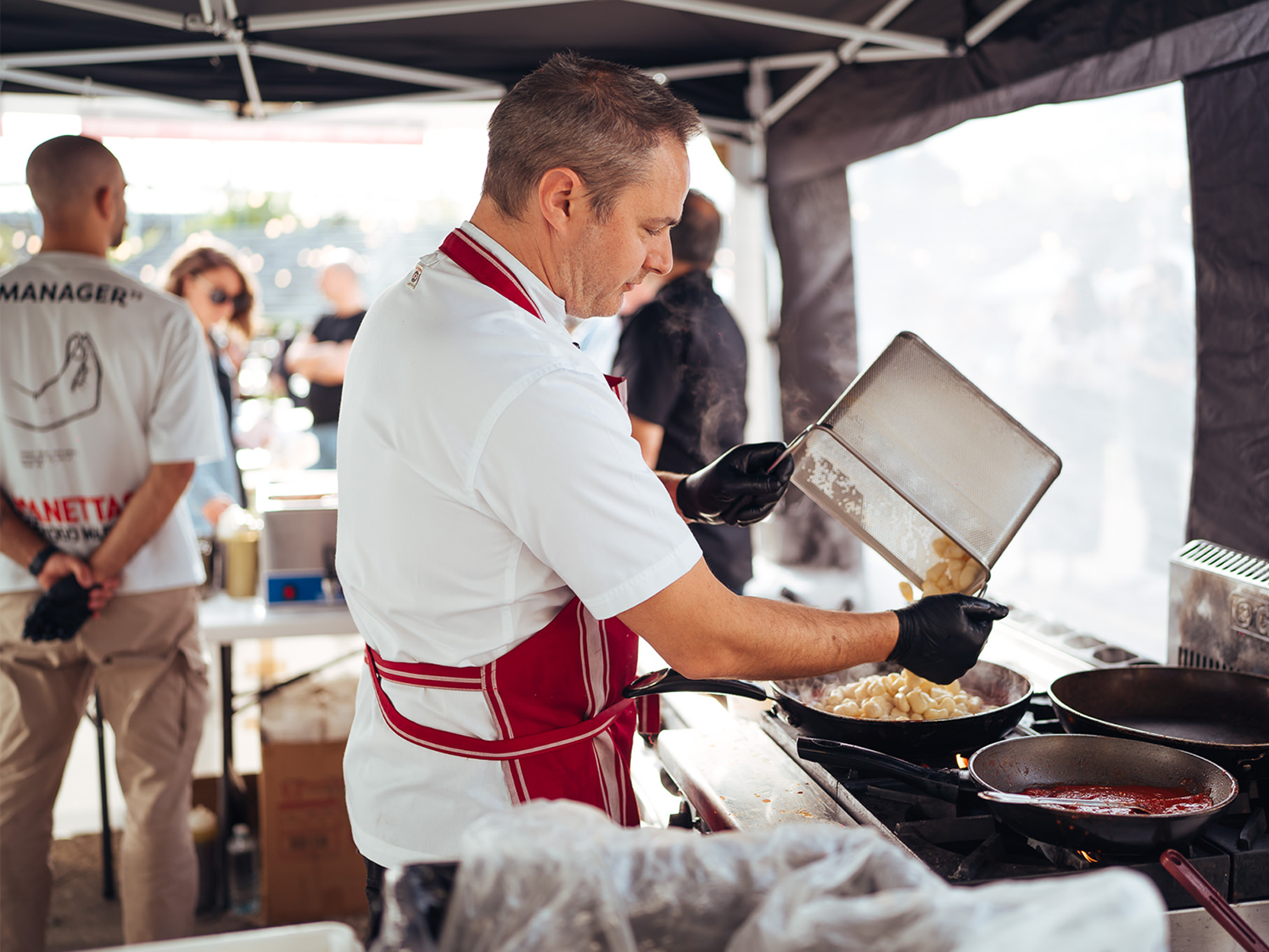 A chef in a red apron cooks food in a frying pan on a stove at an outdoor market stall for Grape, Grain and Graze Festival.