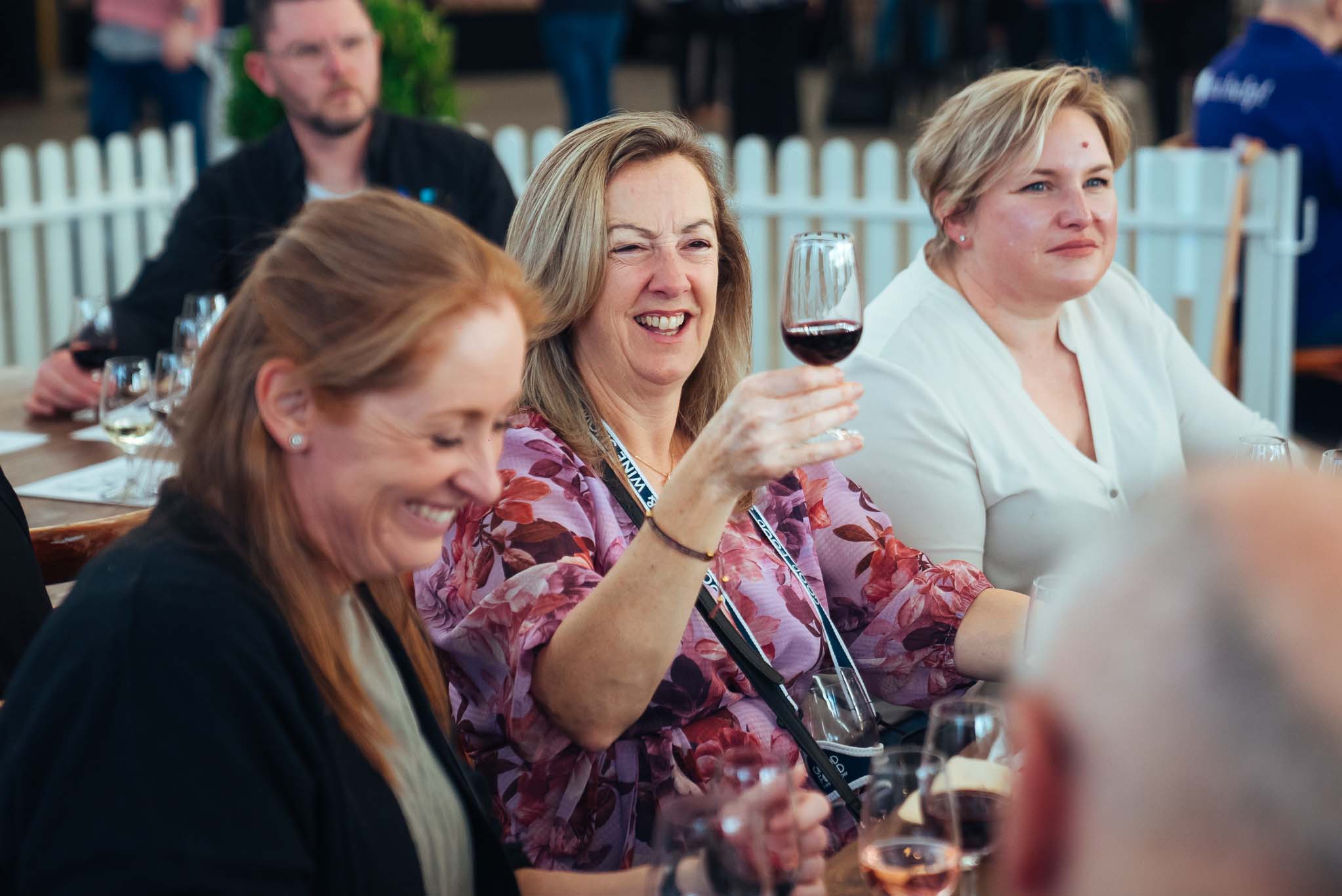 Three women sit at a table enjoying wine tasting at Grape, Grain and Graze Festival, with one woman raising her glass and smiling.