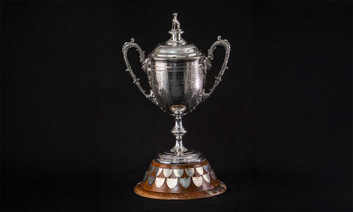 Ornate lidded silver twin handled cup, with tiered domed lid surmounted by a figure of a greyhound, raised on a circular walnut plinth. First presented in 1947. 