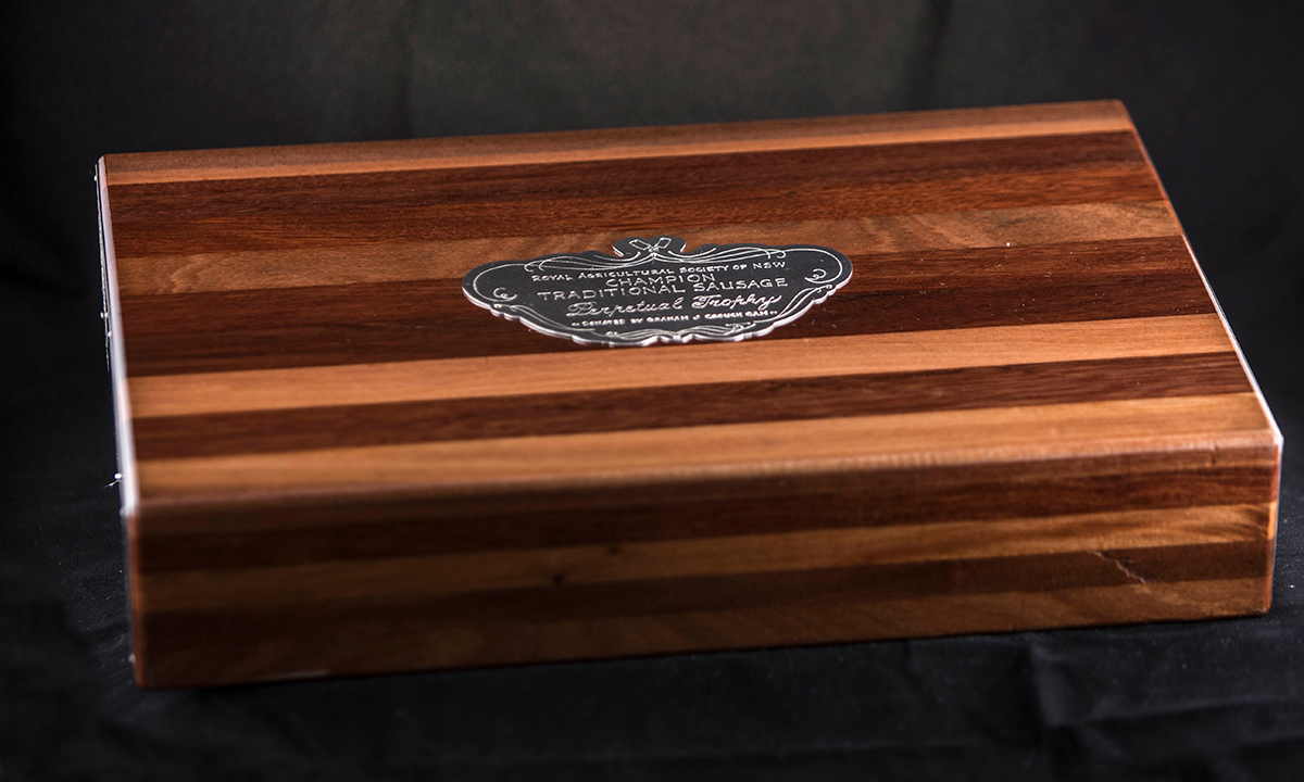 Wooden chopping board with different colours of stained wood used in a stylised block design. Silver name plate on the top. First presented in 2011.
