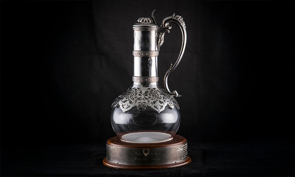 A clear glass wine jug with round body. Decorative silver finishings are used for the neck, spout and handle. The jug is mounted on a circular wooden base. First presented in 2004.