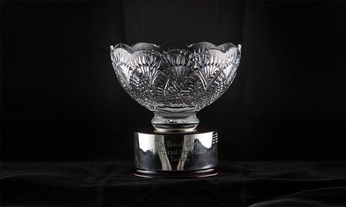 Decorative crystal bowl mounted on a circular wooden base. A silver band wrapped around the base reads: “The David Clarke Memorial Perpetual Trophy”. Awarded since 2012.