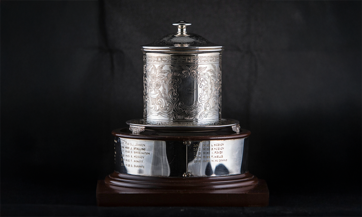 Silver biscuit tin with decorative engraving on the cylinder body and plain lid and finial on top. The tin is on a silver stand, mounted to a circular wooden base with engraving plates. First presented in 1984.