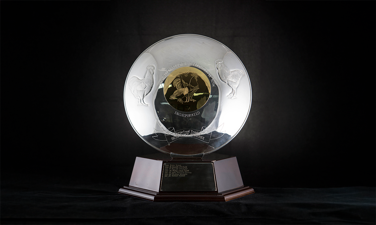 Metal dish featuring decorative pressed metal images of an Australian Langshan and an Australorp either side of a central bronze disk engraved with images of various poultry breeds. A six-sided wooden base supports the dish. First awarded in 2009.