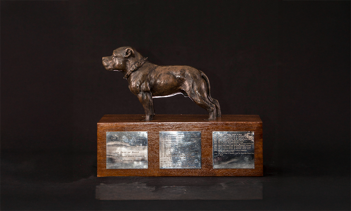 Bronze figure of a Staffordshire Bull Terrier, sitting on top of a rectangular wooden base. First awarded in 1986.