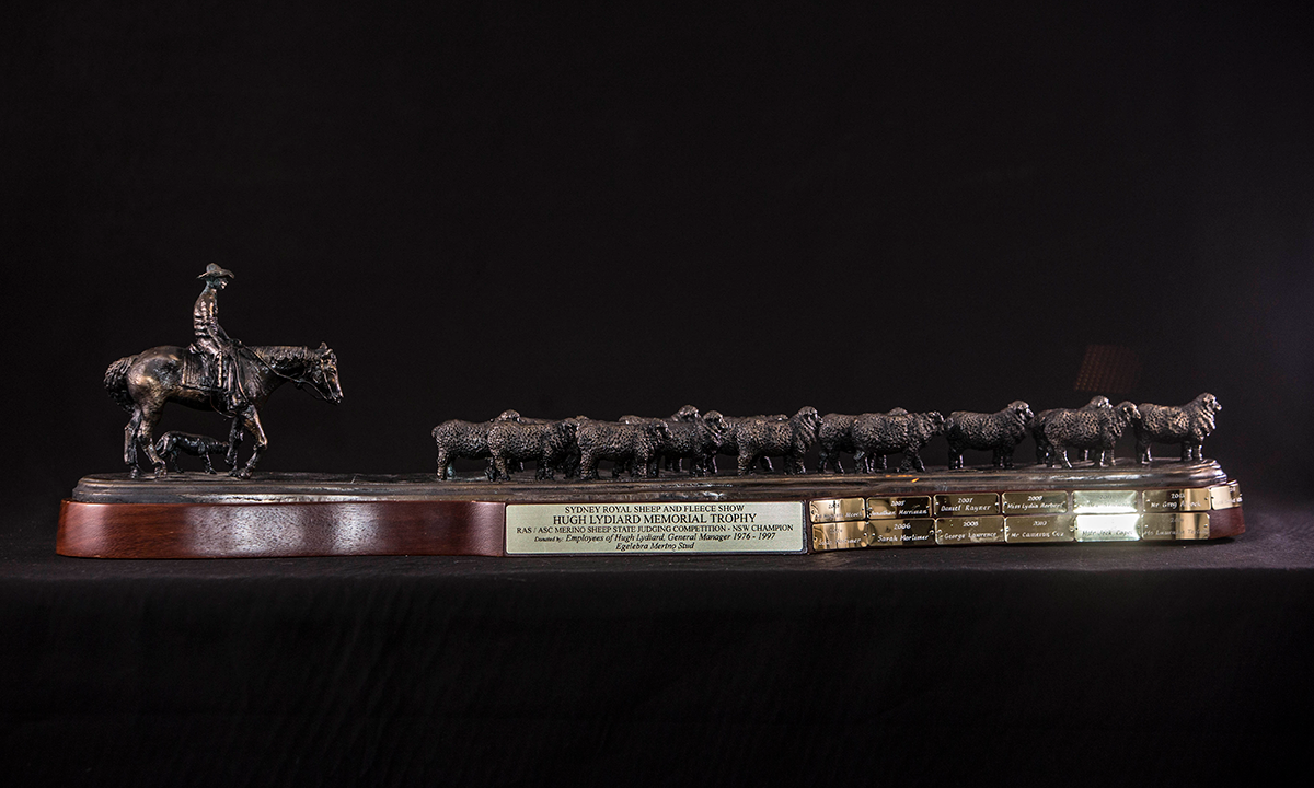 Dark bronze sculpture of a man on his horse with a dog and small flock of sheep, attached to a curved, wooden base with gold engraving plates. First presented in 2003.