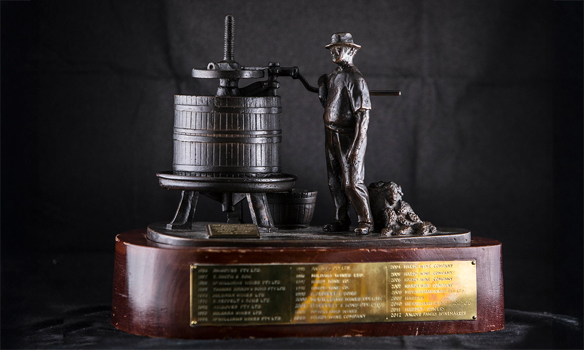 A bronze sculpture by Tom Bass of a vigneron standing beside his grape press, signed and dated '75, raised on an ovoid timber base. First presented in 1986. 