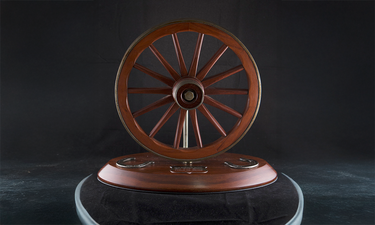 Brass mounted wooden wagon wheel on a base. Two horse-shoe shaped plaques sit either side the trophy name plate on the base. First awarded 1997.