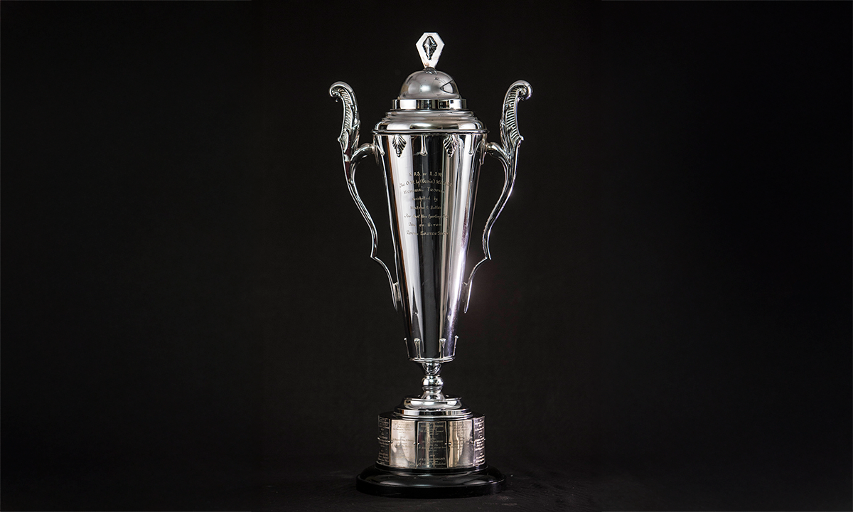 Tall twin handled trophy cup with a cover and diamond-shaped finial, raised on a black composition base. First awarded in 1962.