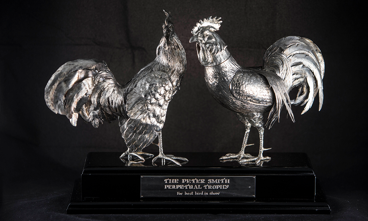Two sterling silver lifelike fighting cocks with detailed feather work face each other, one has its head turned to the side. Both are mounted on a black wooden base. First presented in 2007.
