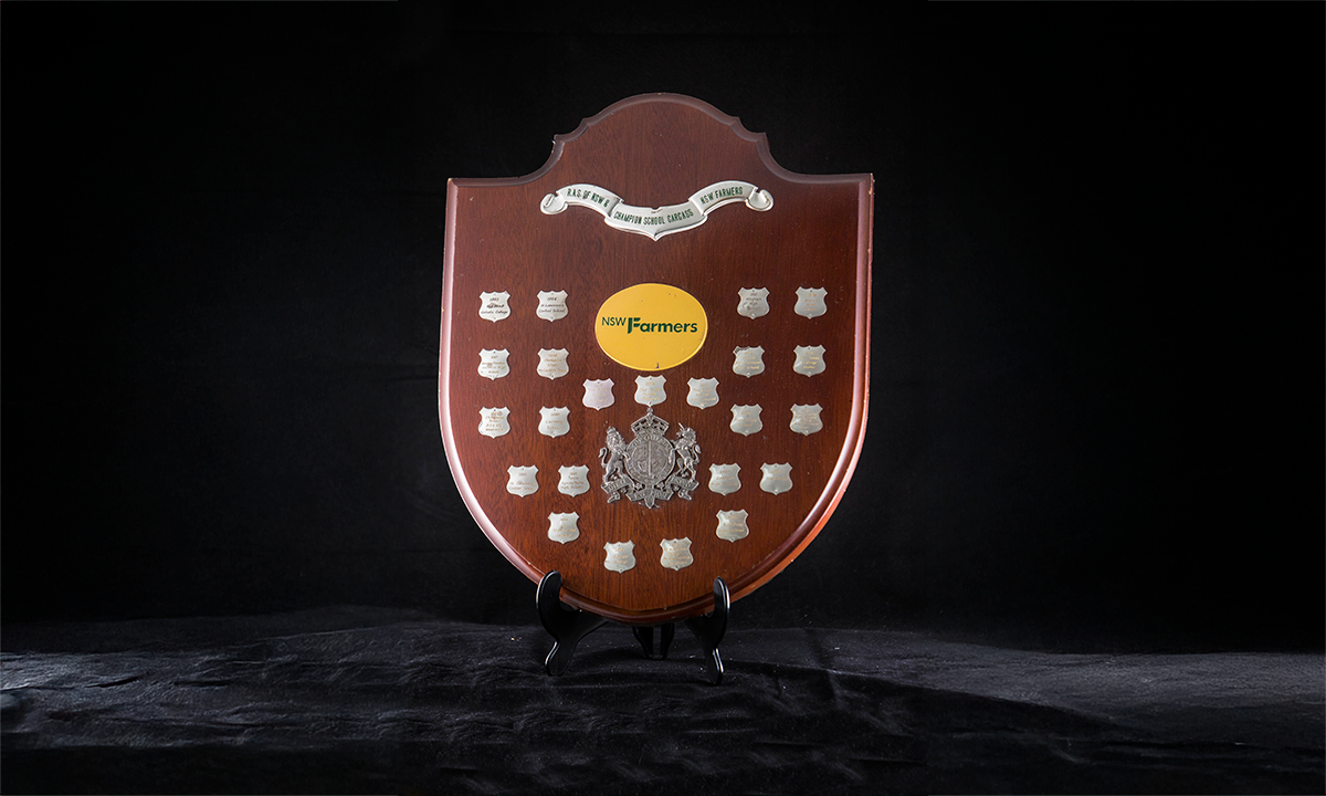 Timber shield with silver plated mounts, a central yellow logo which says ‘NSW Farmers’ and applied coat of arms. First presented in 1993. 