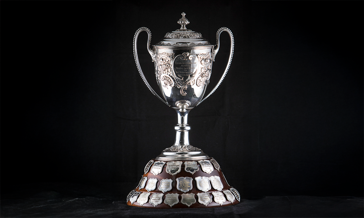 Silver twin handled lidded cup with finial and embossed decoration, raised on a wooden circular plinth. First presented in 1968. 