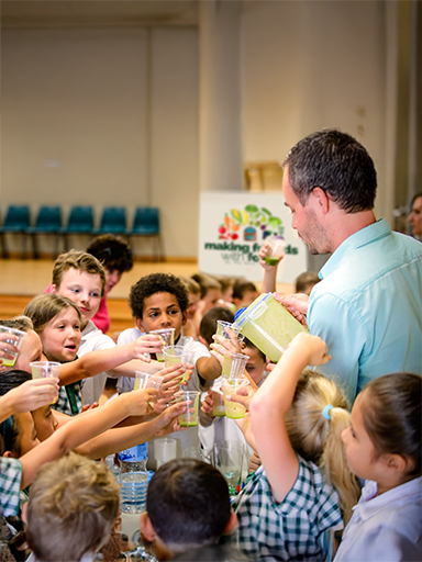 Group of primary school students excitedly holding cups as a man pours green smoothies during a classroom activity.