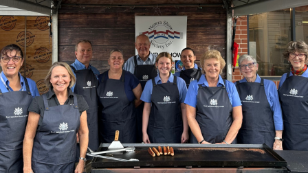 Group of Big Bush BBQ  volunteers wearing navy aprons with Royal Agricultural Society logos stand smiling behind a barbecue at a show stall.