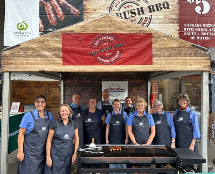 Group of volunteers wearing navy aprons with Royal Agricultural Society logos stand smiling behind a barbecue at a show stall.